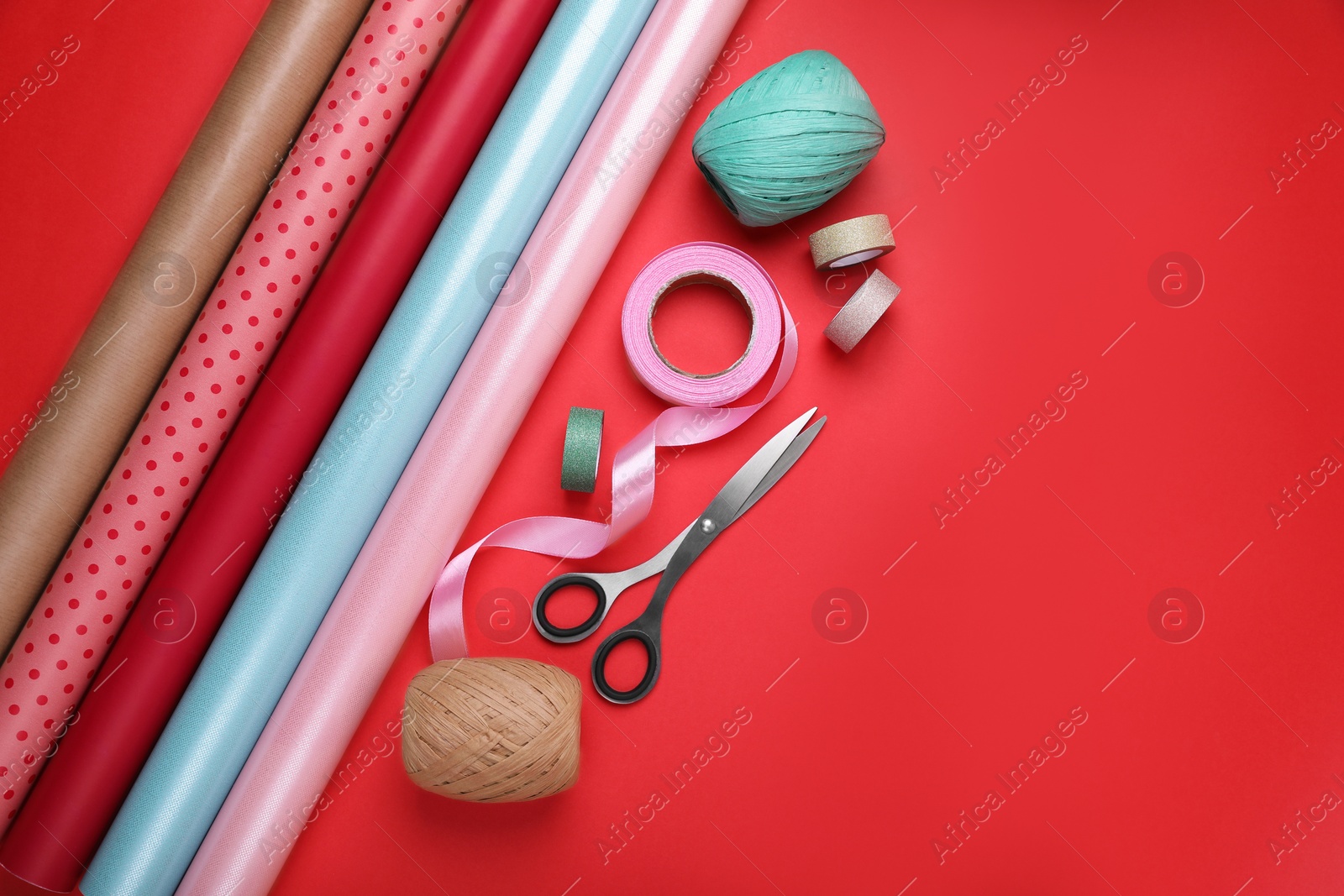 Photo of Rolls of wrapping papers, scissors and ribbons on red background, flat lay. Space for text