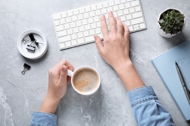 Young woman with cup of coffee and keyboard at grey marble table, top view