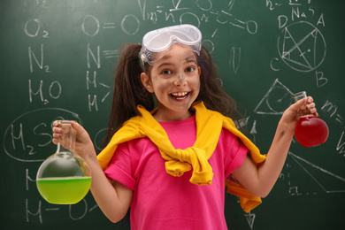 Photo of Schoolgirl holding flasks near chalkboard with chemical formulas