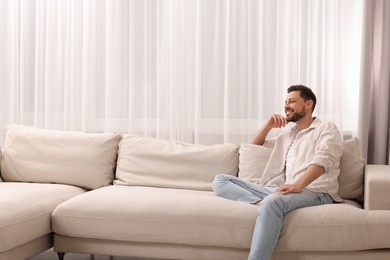 Photo of Happy man on resting sofa near window with beautiful curtains in living room. Space for text