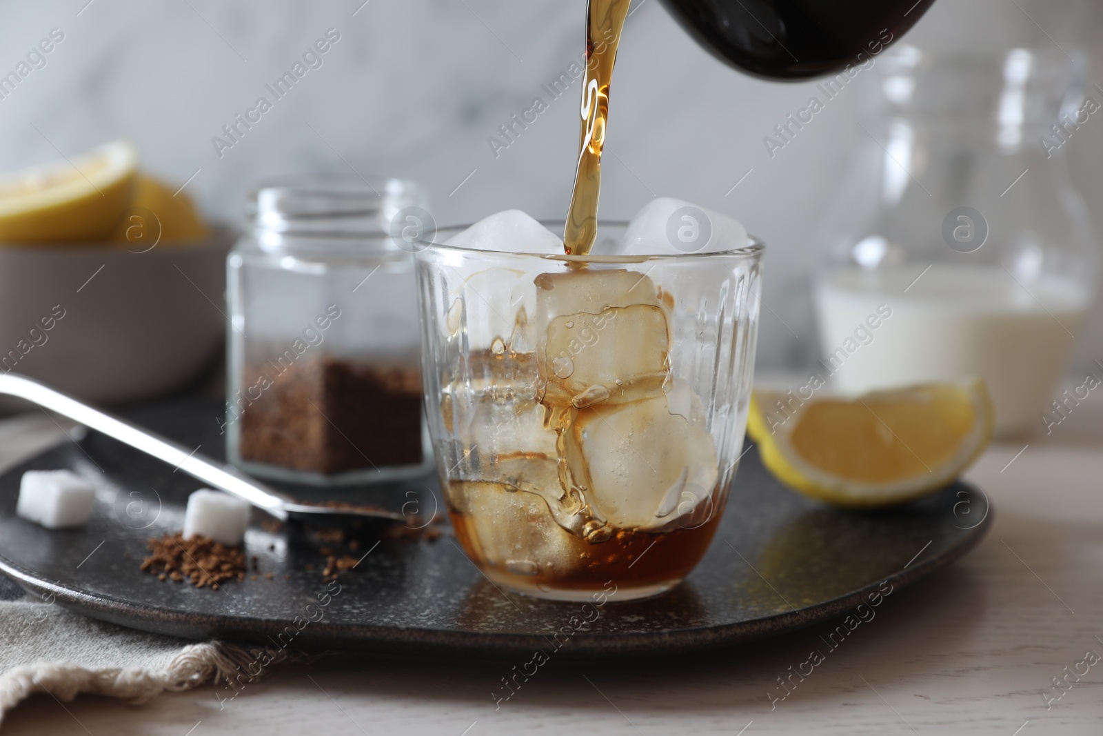 Photo of Pouring coffee into glass with ice cubes at white wooden table, closeup