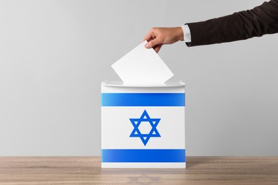 Woman putting her vote into ballot box decorated with flag of Israel against light background, closeup