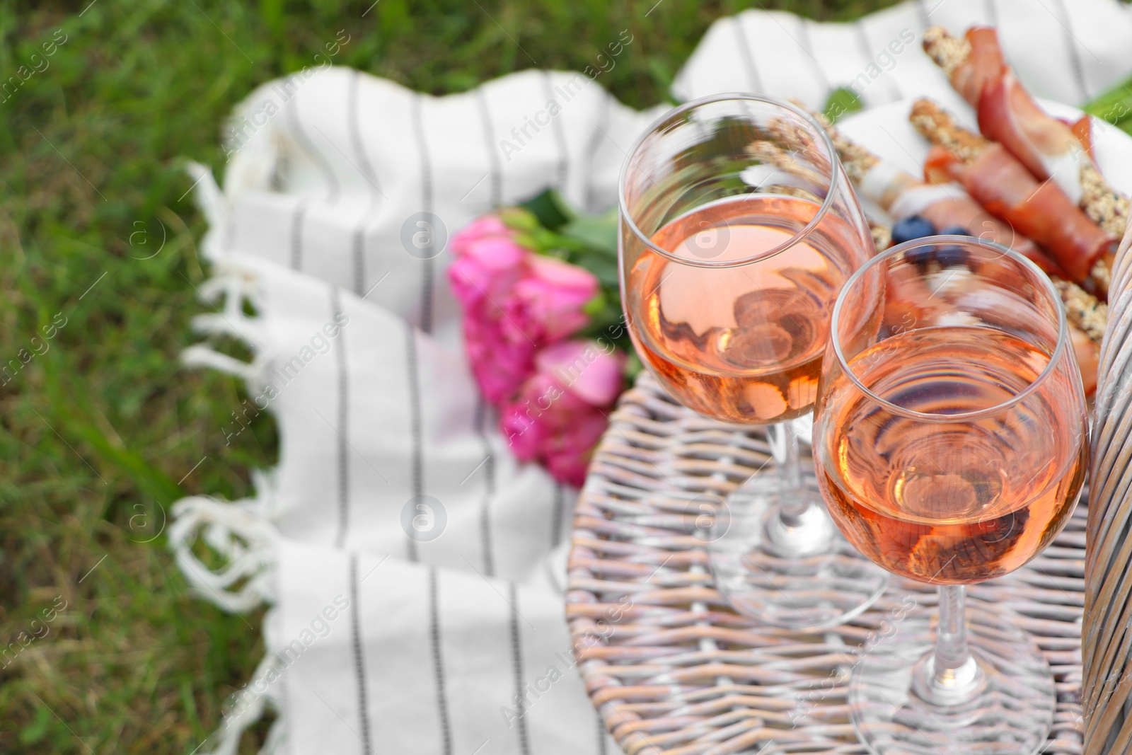 Photo of Glasses of delicious rose wine, food, flowers and basket on picnic blanket outdoors, closeup