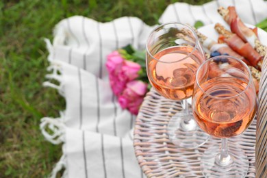 Glasses of delicious rose wine, food, flowers and basket on picnic blanket outdoors, closeup