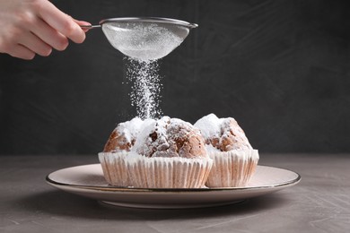 Woman with sieve sprinkling powdered sugar onto muffins at grey textured table, closeup