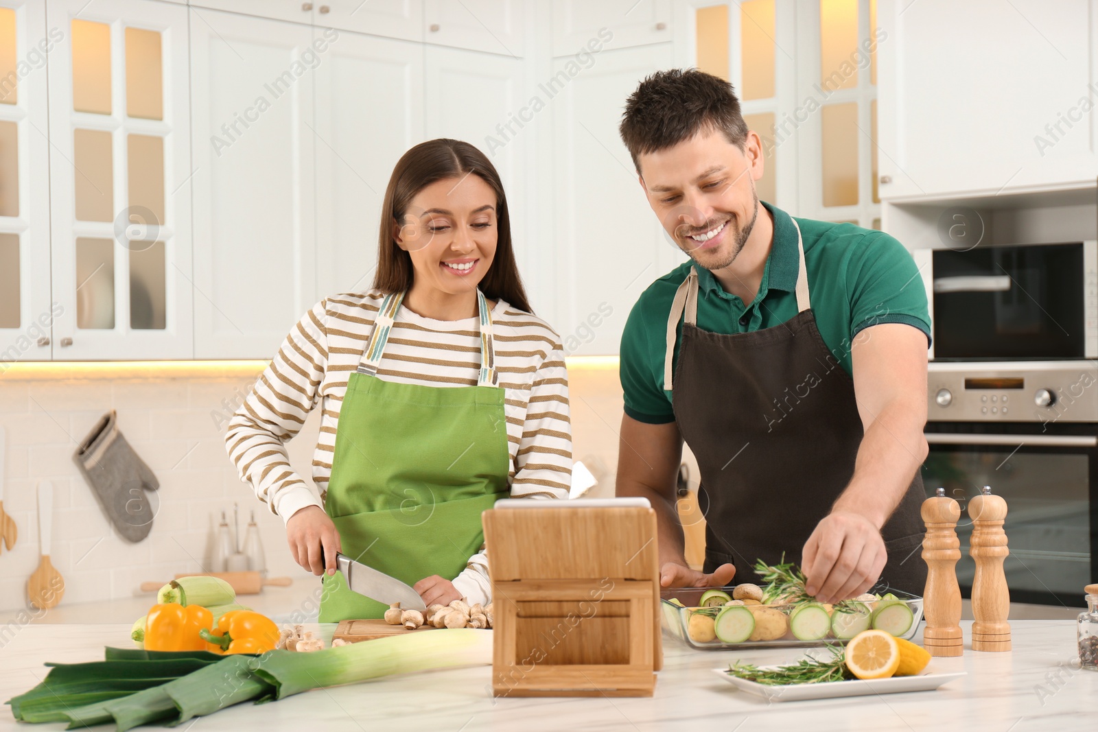 Photo of Happy couple making dinner together while watching online cooking course via tablet in kitchen