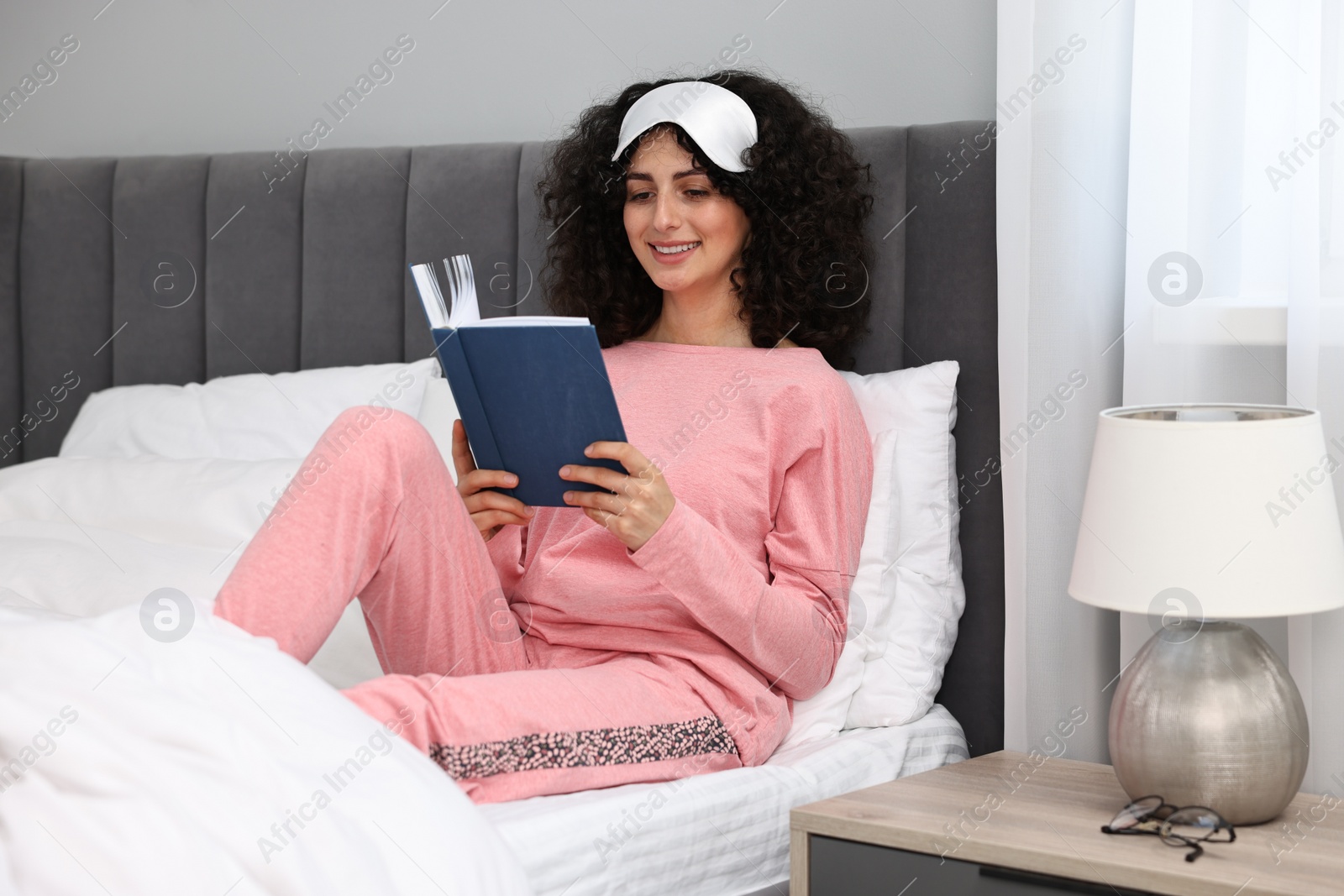 Photo of Beautiful young woman in stylish pyjama and sleep mask reading book on bed at home