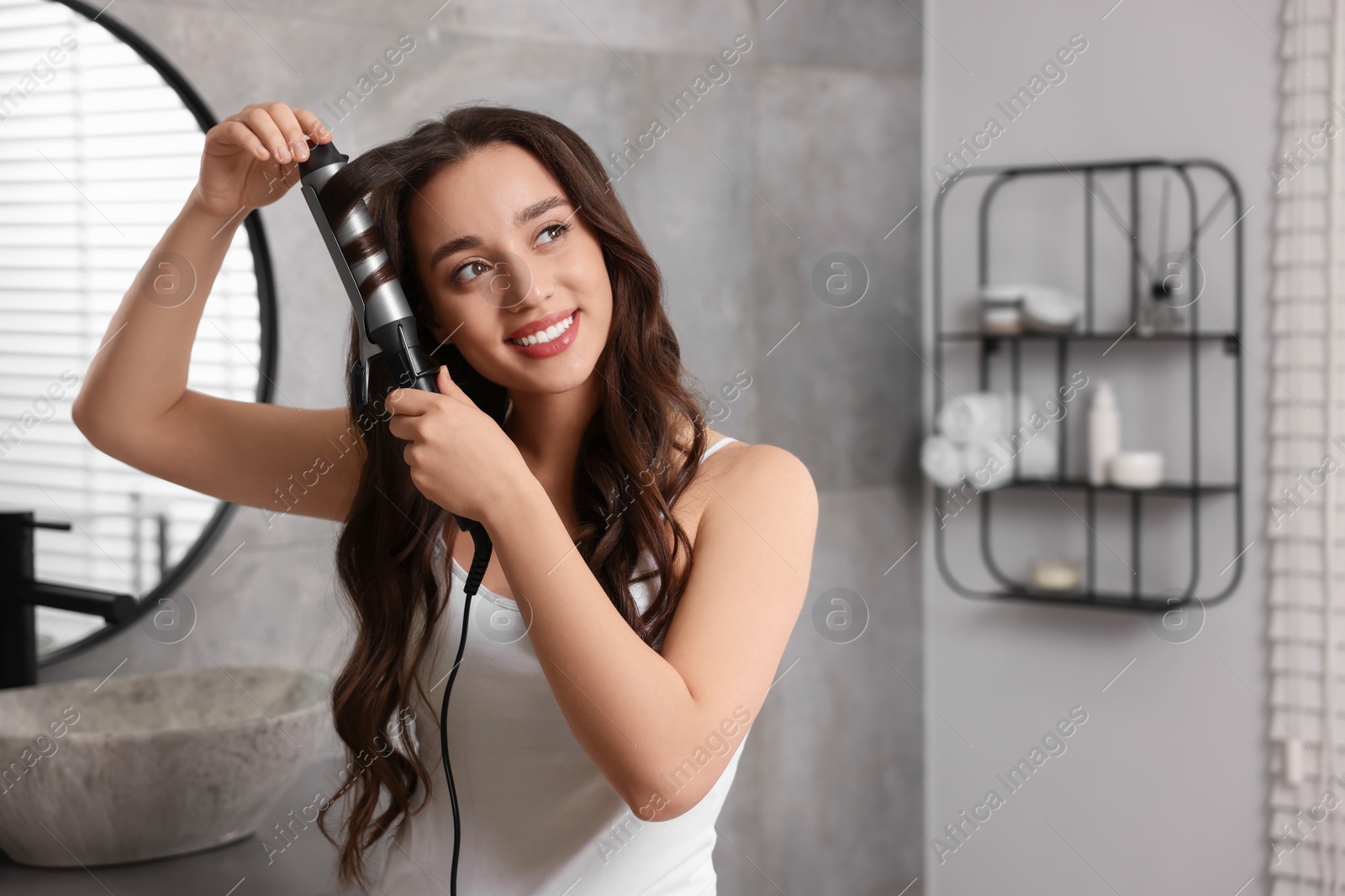 Photo of Smiling woman using curling hair iron in bathroom. Space for text