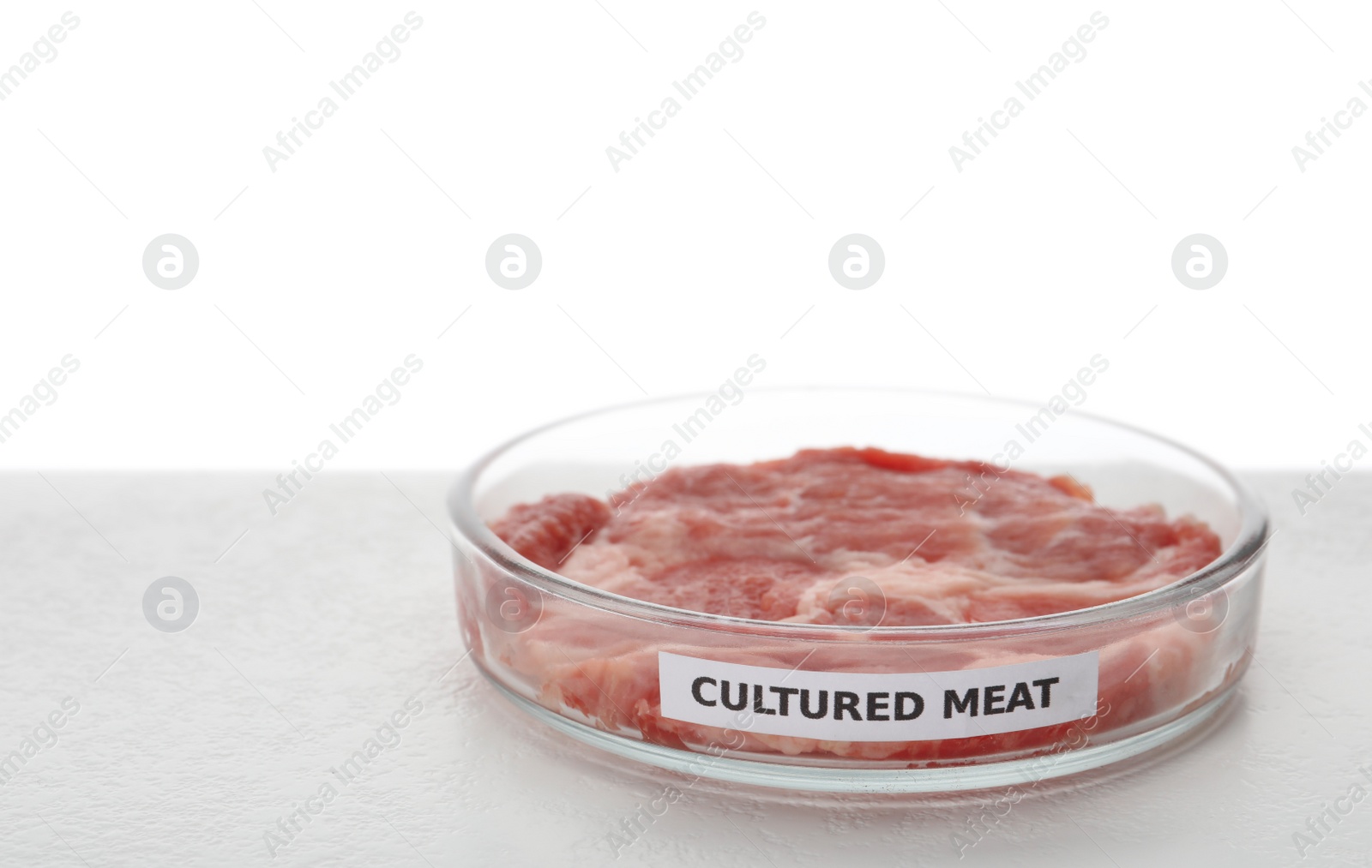 Photo of Petri dish with piece of raw cultured meat and label on table against white background