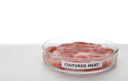 Photo of Petri dish with piece of raw cultured meat and label on table against white background