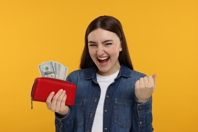 Excited woman holding wallet with dollar banknotes on orange background