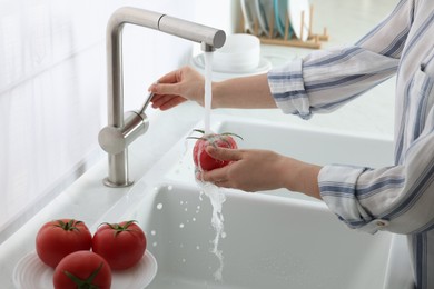 Photo of Woman washing fresh ripe tomato under tap water in kitchen, closeup