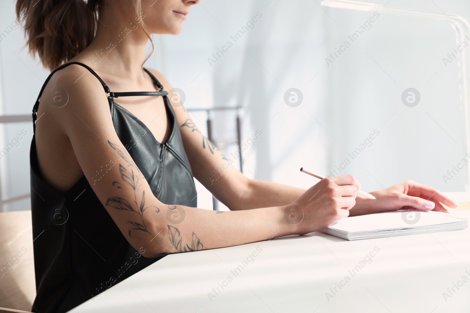 Photo of Beautiful woman with tattoos on body drawing in sketchbook at table indoors, closeup