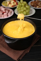 Dipping piece of ham into fondue pot with melted cheese on black wooden table, closeup