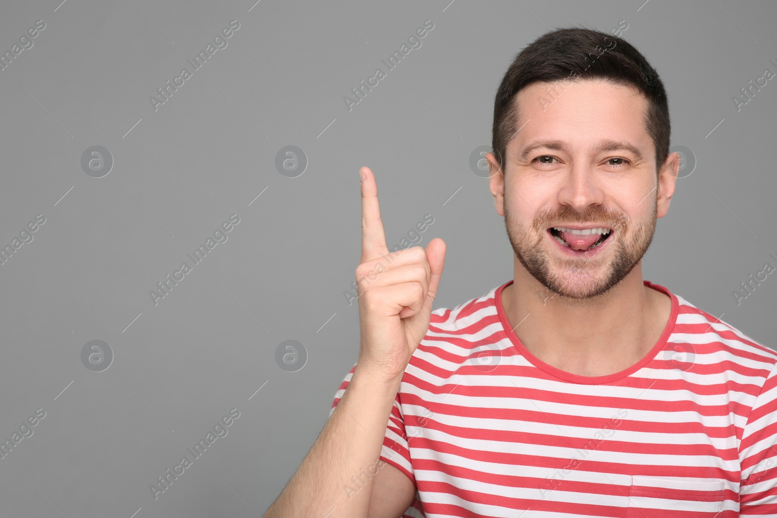 Photo of Happy man showing his tongue and pointing at something on grey background. Space for text