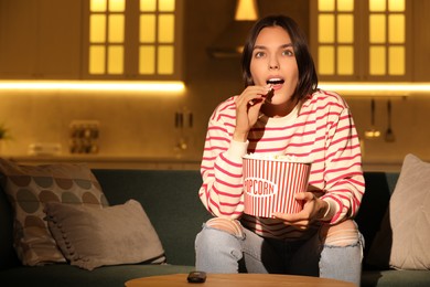 Surprised woman eating popcorn while watching TV on sofa at home, space for text