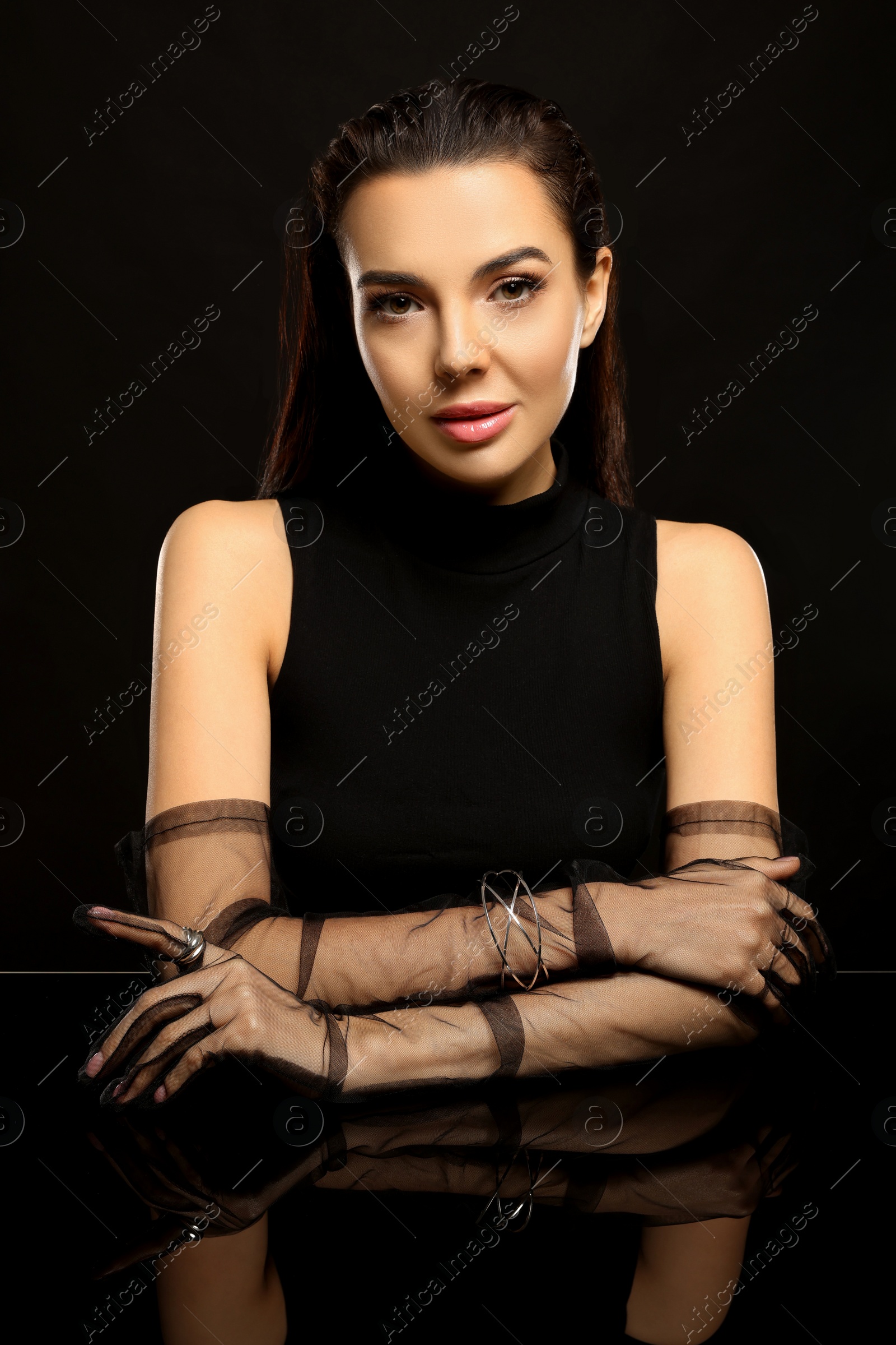 Photo of Beautiful young woman in evening gloves at black glass table against dark background