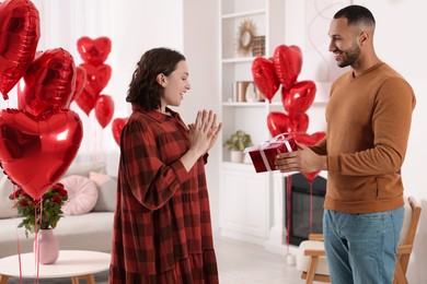 Photo of Boyfriend presenting gift to his girlfriend in room decorated with heart shaped air balloons. Valentine's day celebration