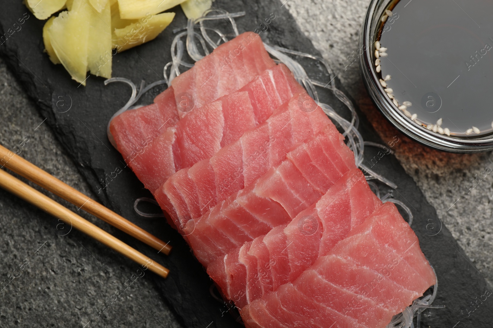 Photo of Tasty sashimi (pieces of fresh raw tuna), glass noodles, soy sauce and chopsticks on gray table, flat lay