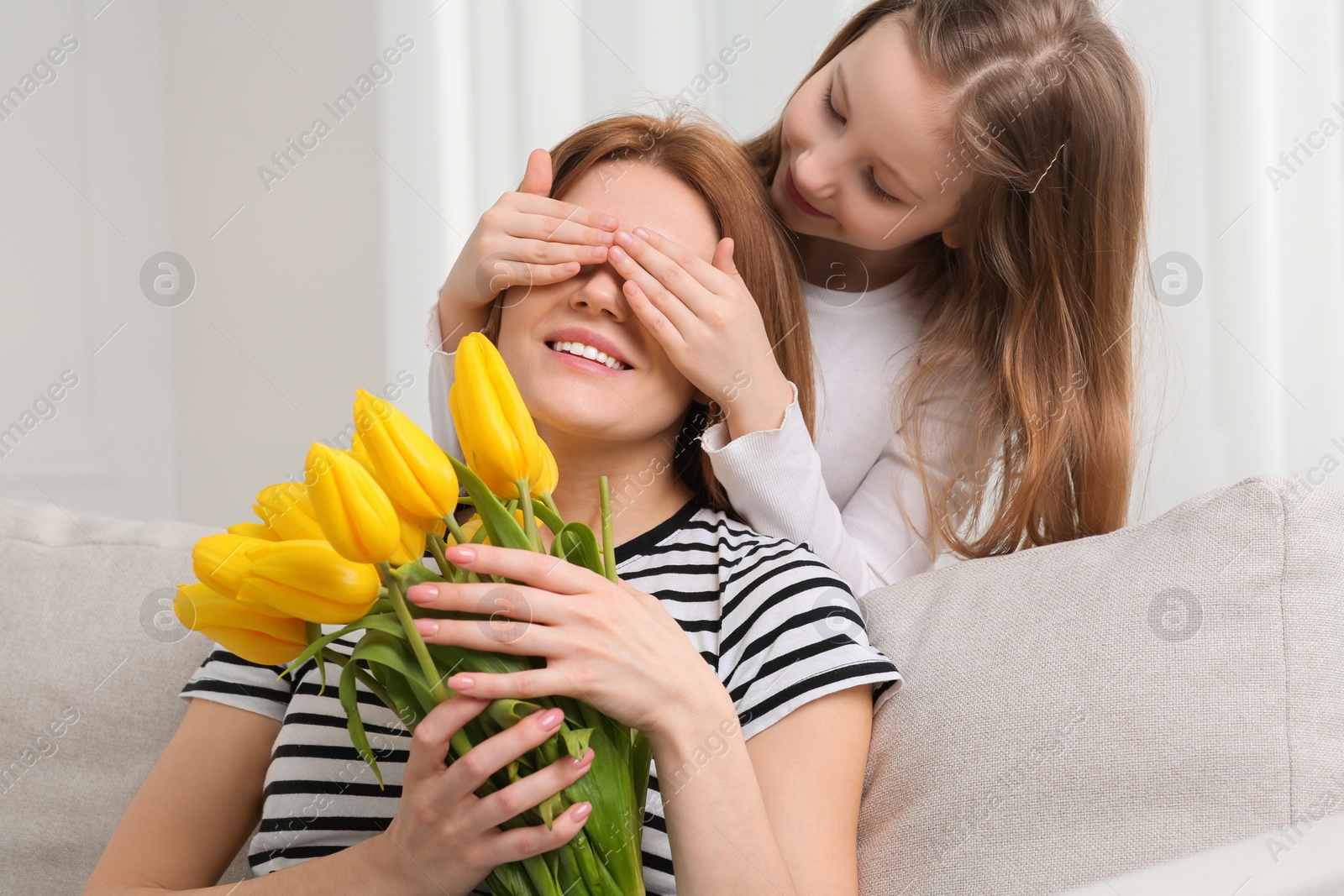 Photo of Daughter covering mother's eyes with her palms and congratulating with bouquet of yellow tulips at home