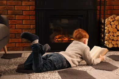 Photo of Boy reading book on floor near fireplace at home, back view