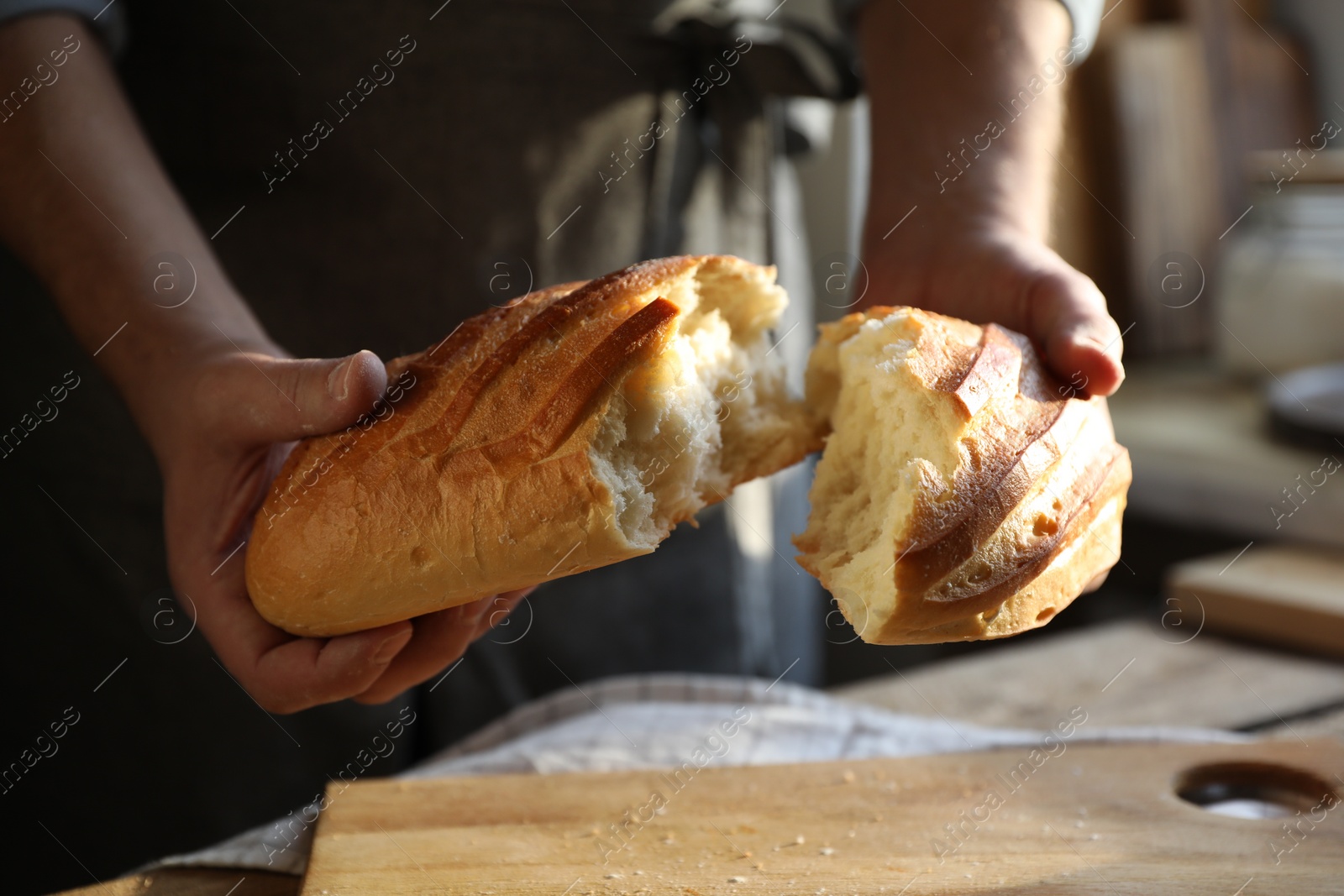 Photo of Man breaking loaf of fresh bread at wooden table indoors, closeup