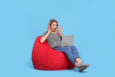 Emotional woman with laptop sitting on beanbag chair against light blue background