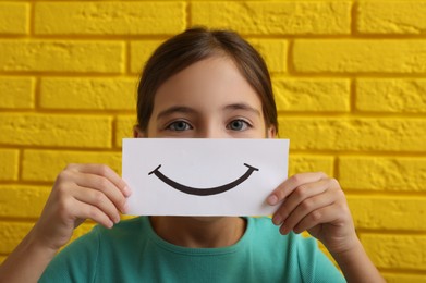 Photo of Little girl holding sheet of paper with smile against yellow brick wall