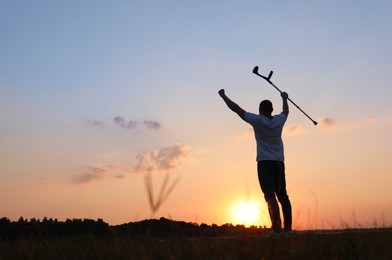 Photo of Man raising elbow crutch up to sky outdoors at sunset, back view and space for text. Healing miracle