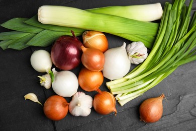 Photo of Fresh onion bulbs, leeks and garlic on black table, flat lay