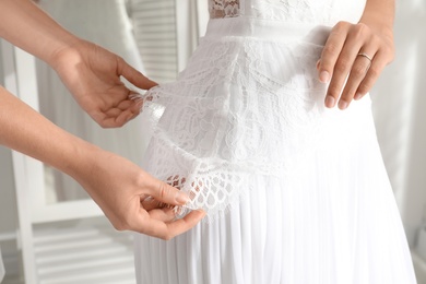 Woman helping bride to put on wedding dress indoors, closeup