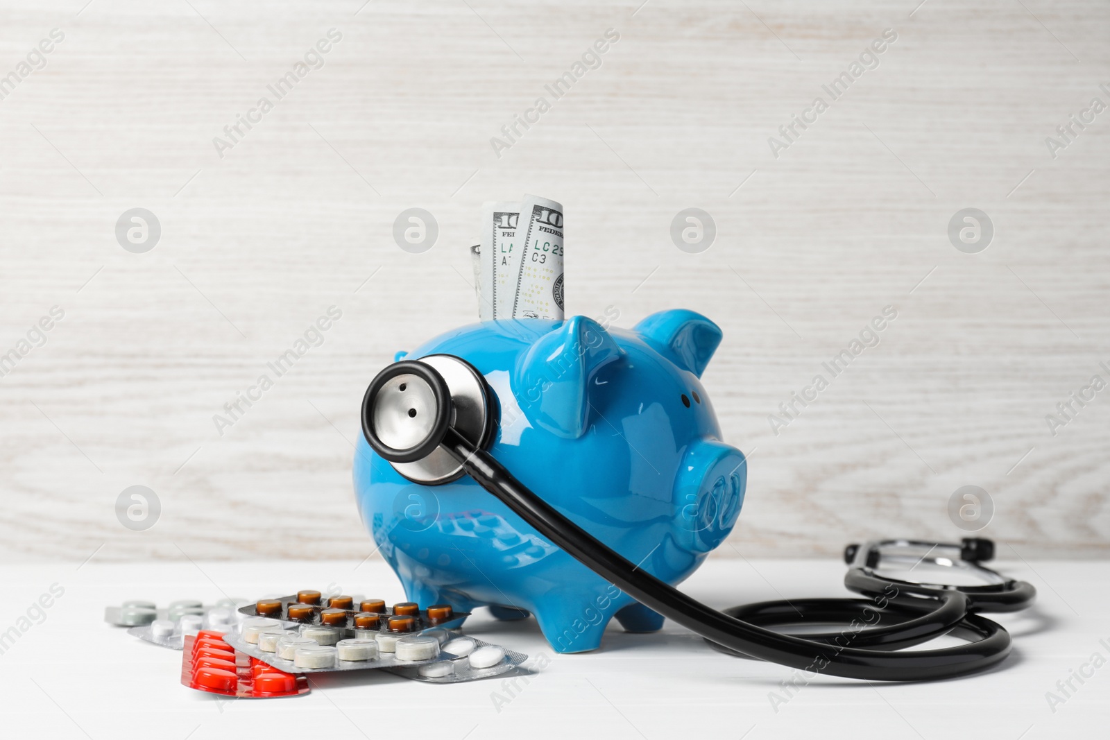 Photo of Light blue ceramic piggy bank with money, stethoscope and pills on white wooden table. Medical insurance