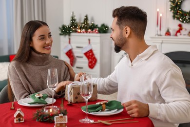 Happy young man presenting Christmas gift to his girlfriend at table indoors