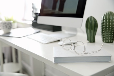 Photo of Comfortable workplace with computer on desk in home office, focus on book and glasses