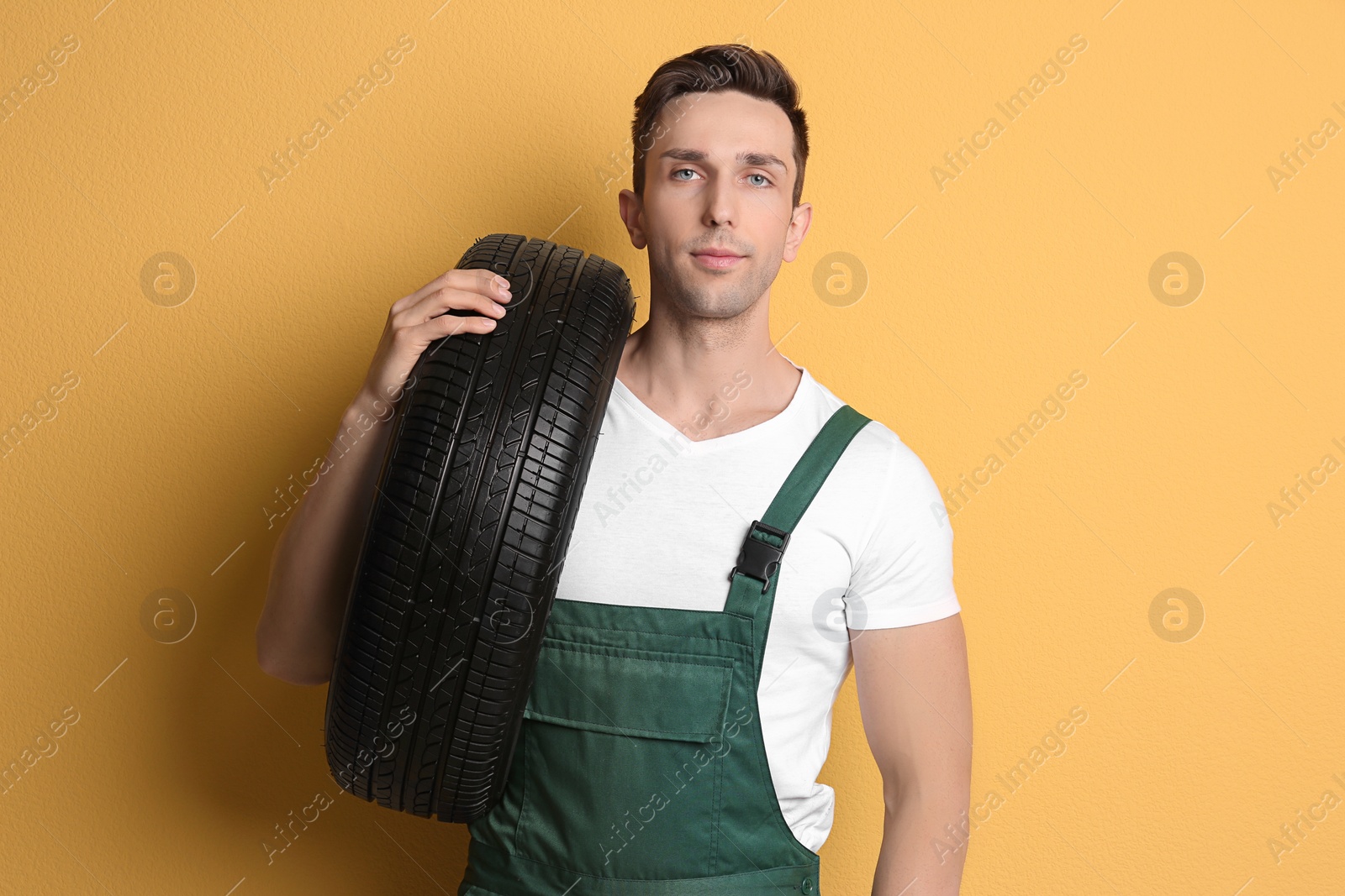 Photo of Young mechanic in uniform holding car tire on color background