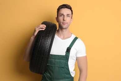 Young mechanic in uniform holding car tire on color background