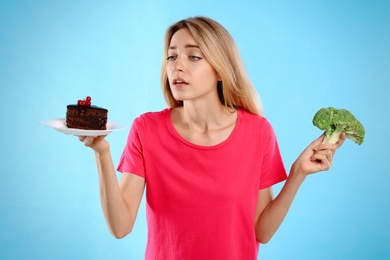 Woman choosing between cake and healthy broccoli on light blue background