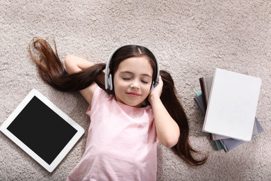 Cute little girl with headphones and tablet listening to audiobook on floor indoors, flat lay