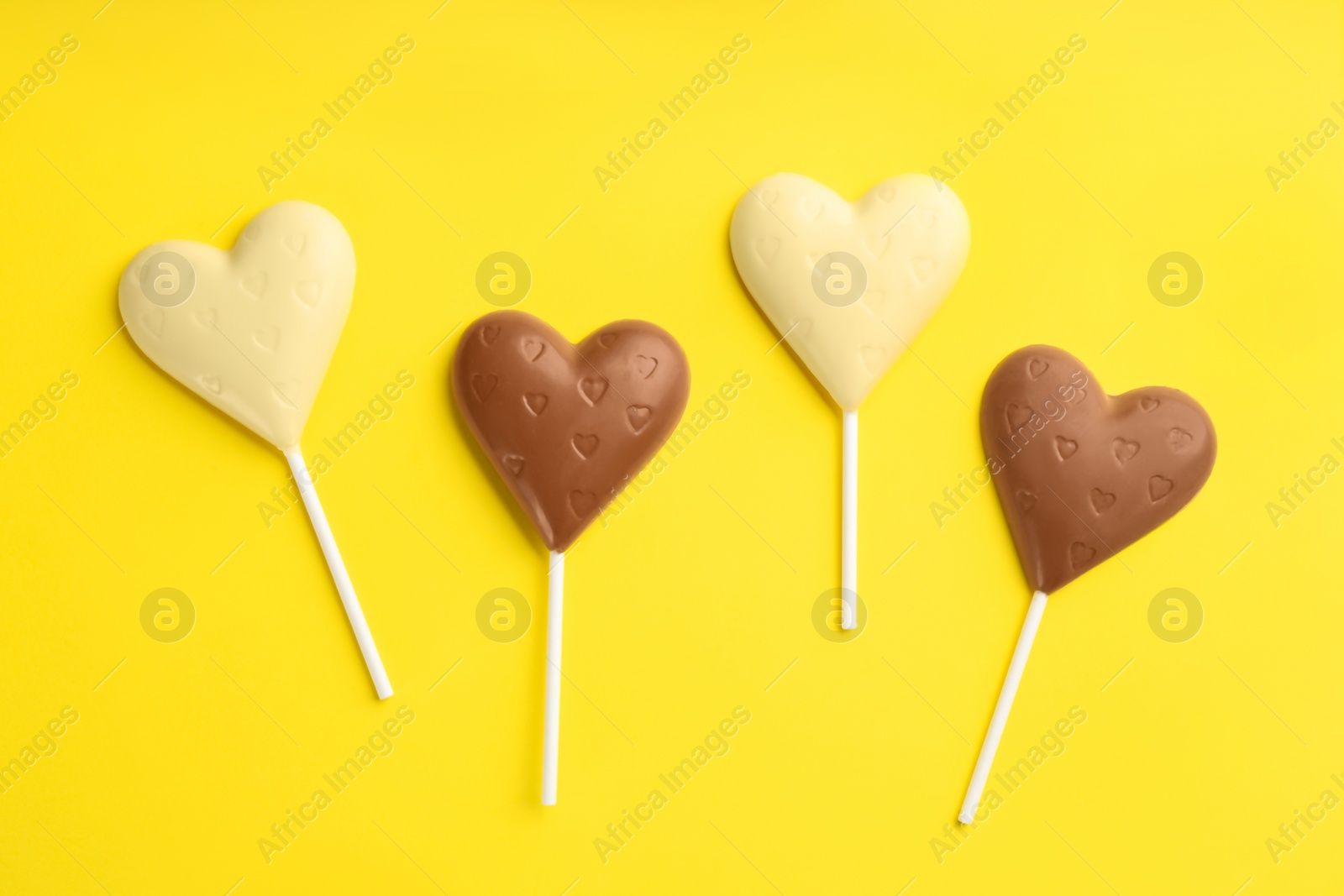 Photo of Different chocolate heart shaped lollipops on yellow background, flat lay