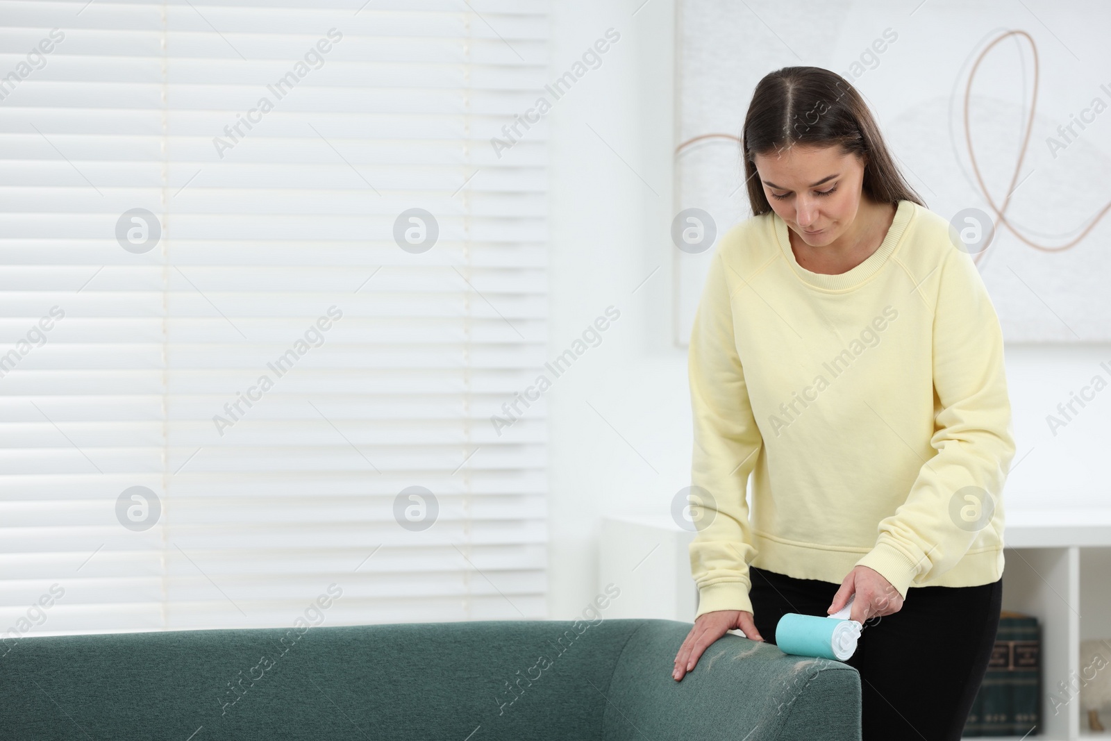 Photo of Woman with lint roller removing pet hair from sofa at home, space for text