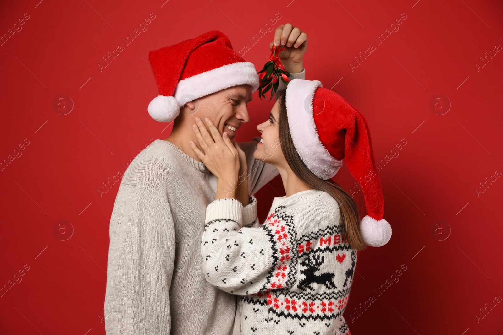 Photo of Happy couple in Santa hats standing under mistletoe bunch on red background