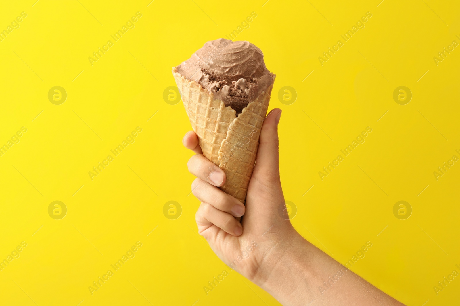 Photo of Woman holding waffle cone with delicious chocolate ice cream on yellow background, closeup
