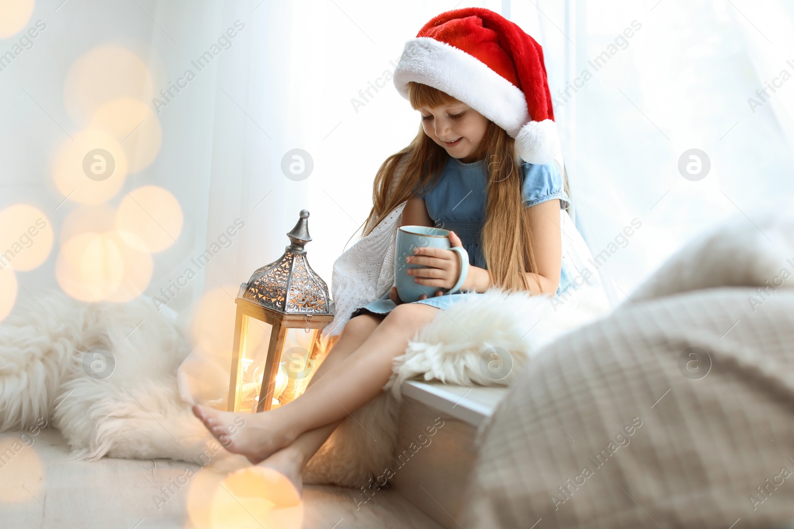 Photo of Cute little child in Santa hat with cup of cocoa sitting on windowsill at home. Christmas celebration