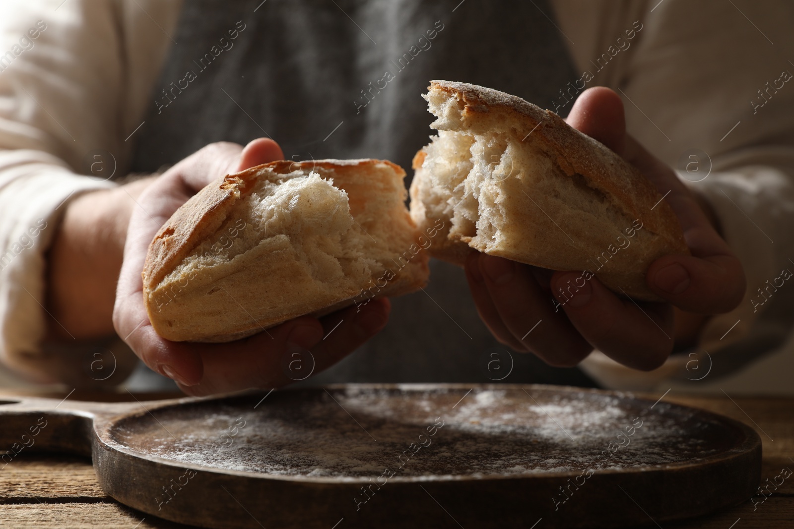 Photo of Man breaking loaf of fresh bread at wooden table, closeup