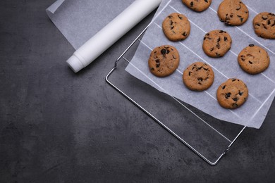 Photo of Roll of parchment baking paper and cooling rack with tasty cookies on dark grey table, flat lay. Space for text