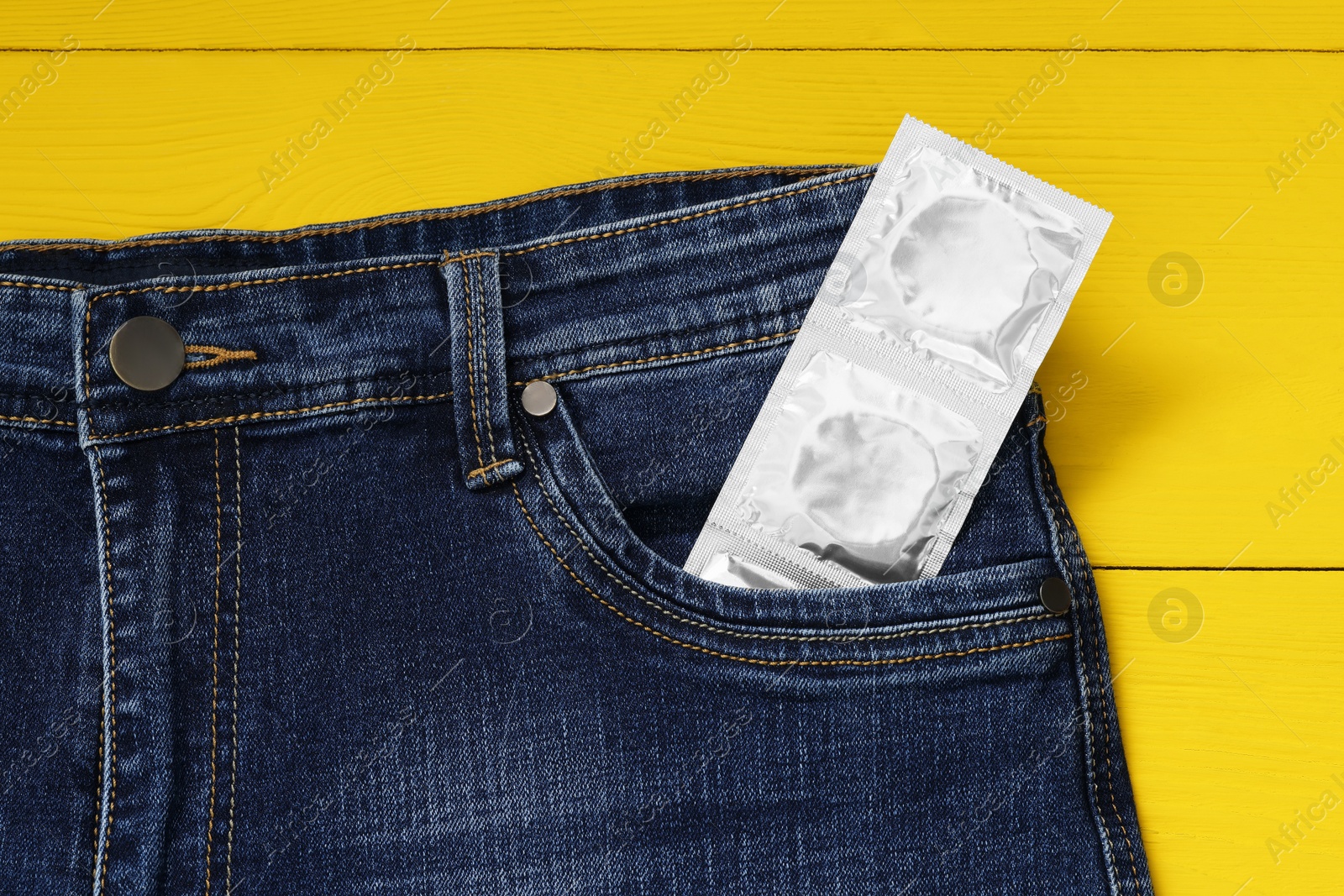 Photo of Packaged condoms in jeans pocket on yellow wooden table, top view. Safe sex