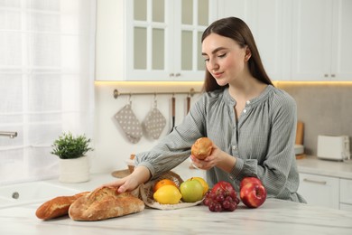 Photo of Woman with baguette, bread and string bag of fresh fruits at light marble table in kitchen