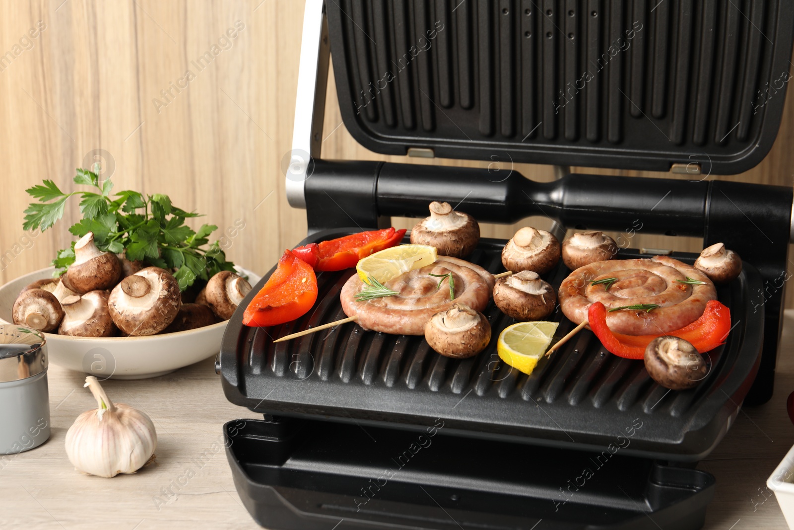 Photo of Electric grill with homemade sausages, vegetables and spices on wooden table