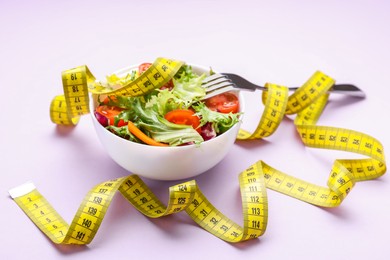 Photo of Bowl with fresh vegetable salad, fork and measuring tape on violet background. Healthy diet concept