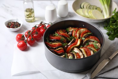 Photo of Delicious ratatouille in round baking pan, knife and ingredients on white table